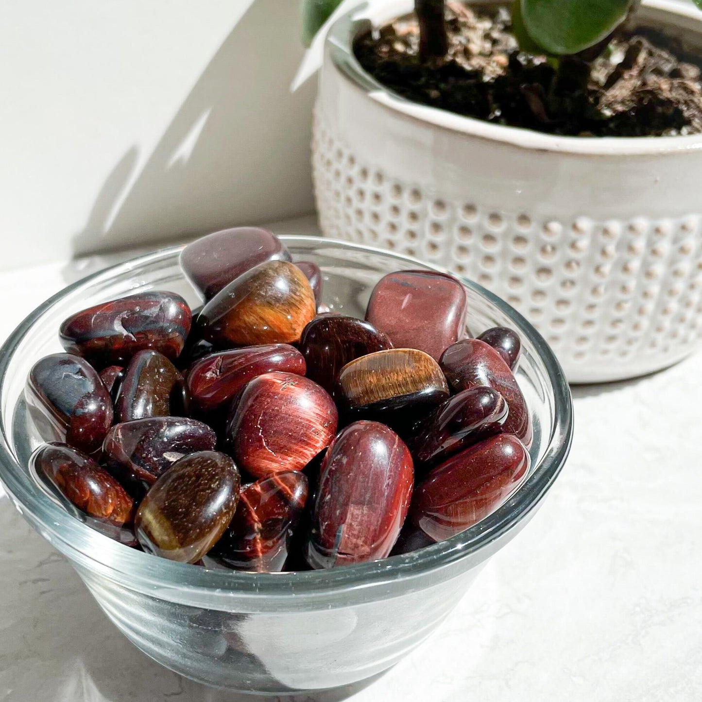 A collection of red tiger eye stones in a clear glass bowl, with a plant pot in the background.