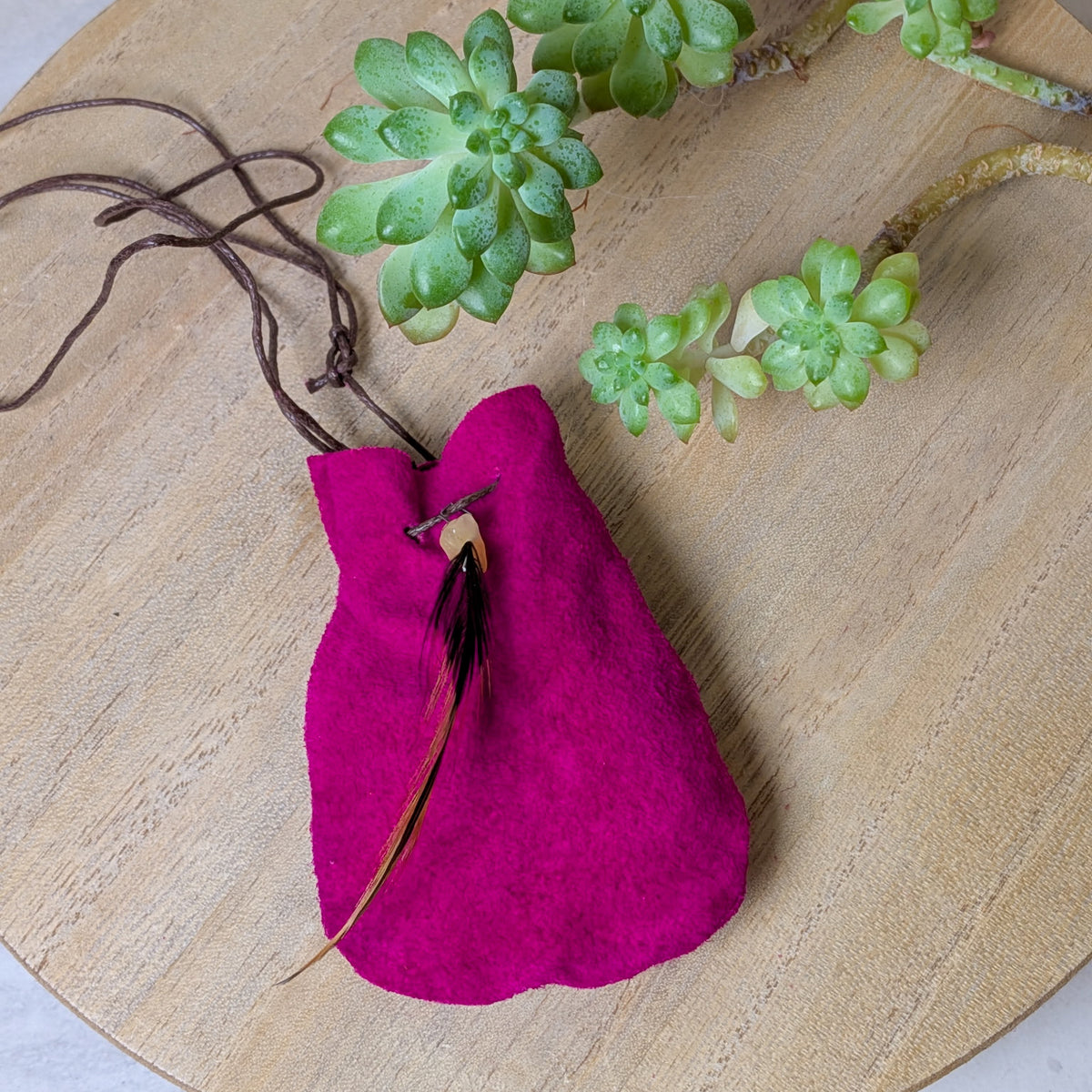 A small magenta fabric pouch with a brown drawstring and a feather accent, styled on a light wood surface with green succulent plants.
