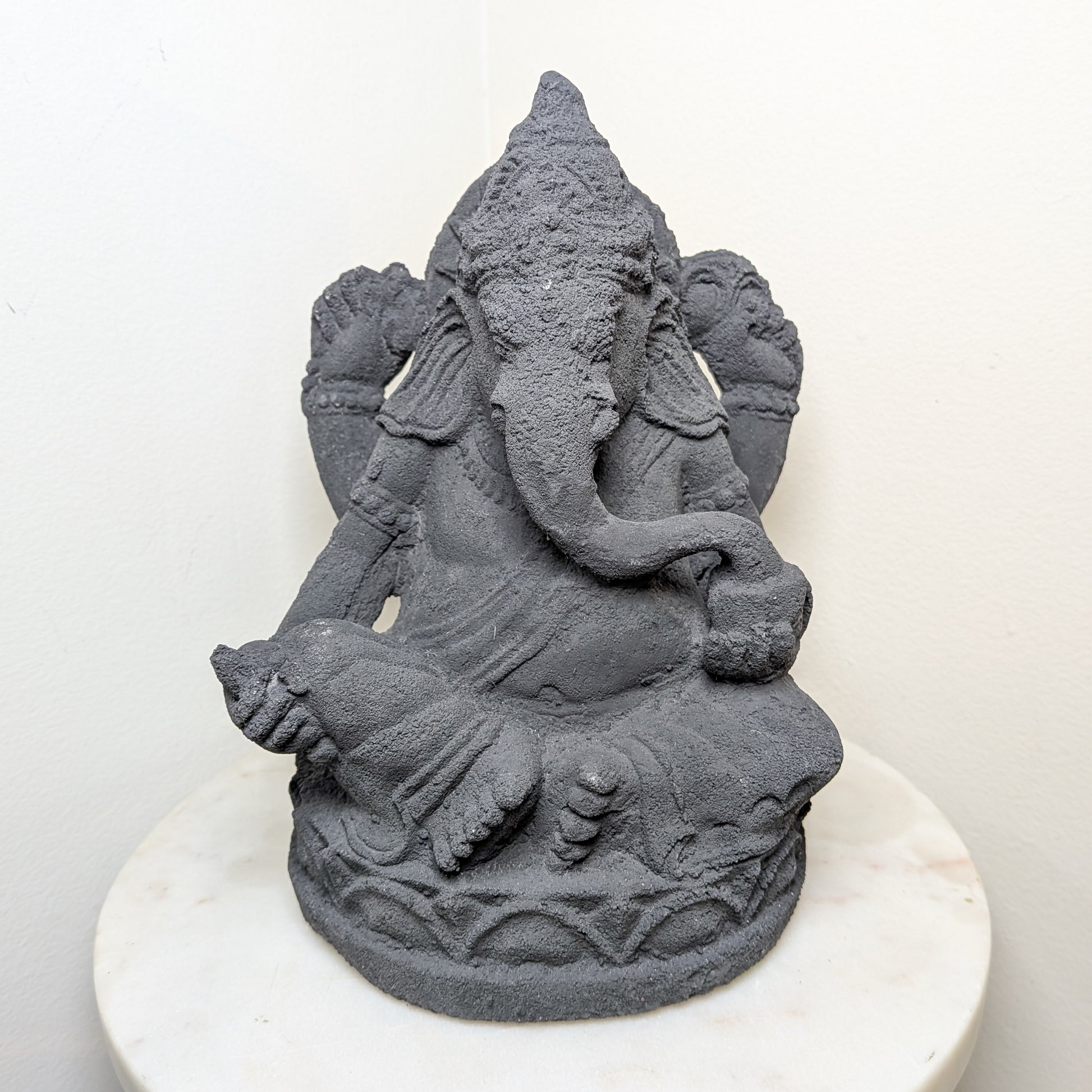 Gray stone statue of an elephant headed deity named Ganesh on a white pedestal against a white background.