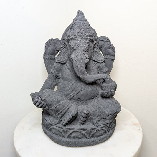 Gray stone statue of an elephant headed deity named Ganesh on a white pedestal against a white background.
