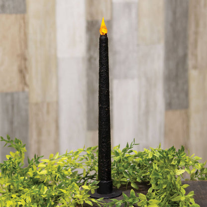 A black textured taper candle with a glowing orange flame, displayed in a black holder surrounded by green foliage against a blurred wood-paneled background.